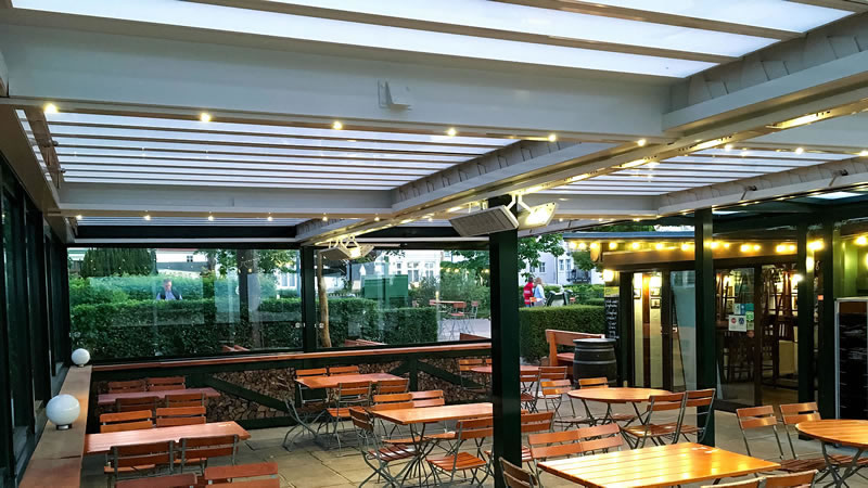 Innenansicht - Restaurantterrasse mit Lamellendach und seitlicher verschiebbarer Verglasung als  Wind- und Wetterschutz.