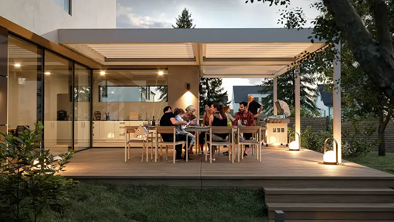 FLEDMEX<sup>®</sup> Lamellendach als stimmungsvolle Terrassenüberdachung beim gemeinsamen Essen und Grillen an einem Sommerabend.