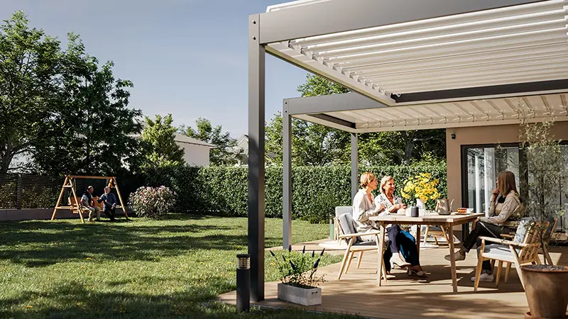 FLEDMEX<sup>®</sup> Lamellendach als elegante Terrassenüberdachung im sonnigen Garten, während mehrere Personen am Tisch sitzen und weitere Personen im Hintergrund die Zeit im Freien genießen.